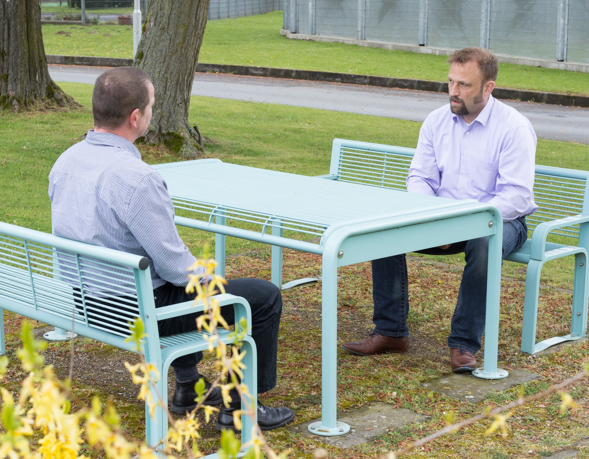 Zwei Männer sitzen an einem Tisch im Freien und unterhalten sich. Im Hintergrund steht ein Sicherheitszaun in Höhe von 5,50 Metern.
