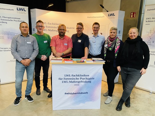 Fünf Männer und zwei Frauen stehen an einem Infostand mit der Aufschrift "LWL-Maßregelvollzug" und lächeln in die Kamera (Foto: LWL/Hekel)