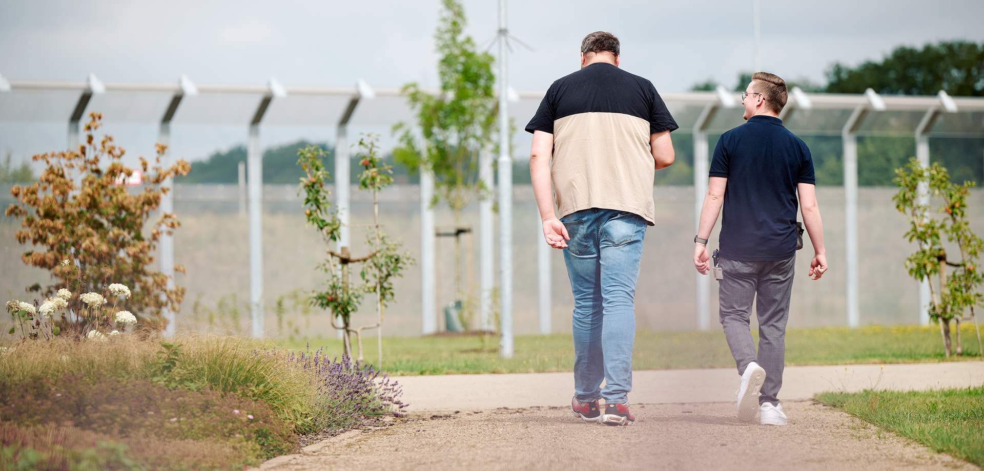 Zwei Männer gehen einen Weg entlang. Man sieht sie nur von hinten. Im Hindergrund befindet sich ein durchsichtiger hoher Sicherheitszaun.